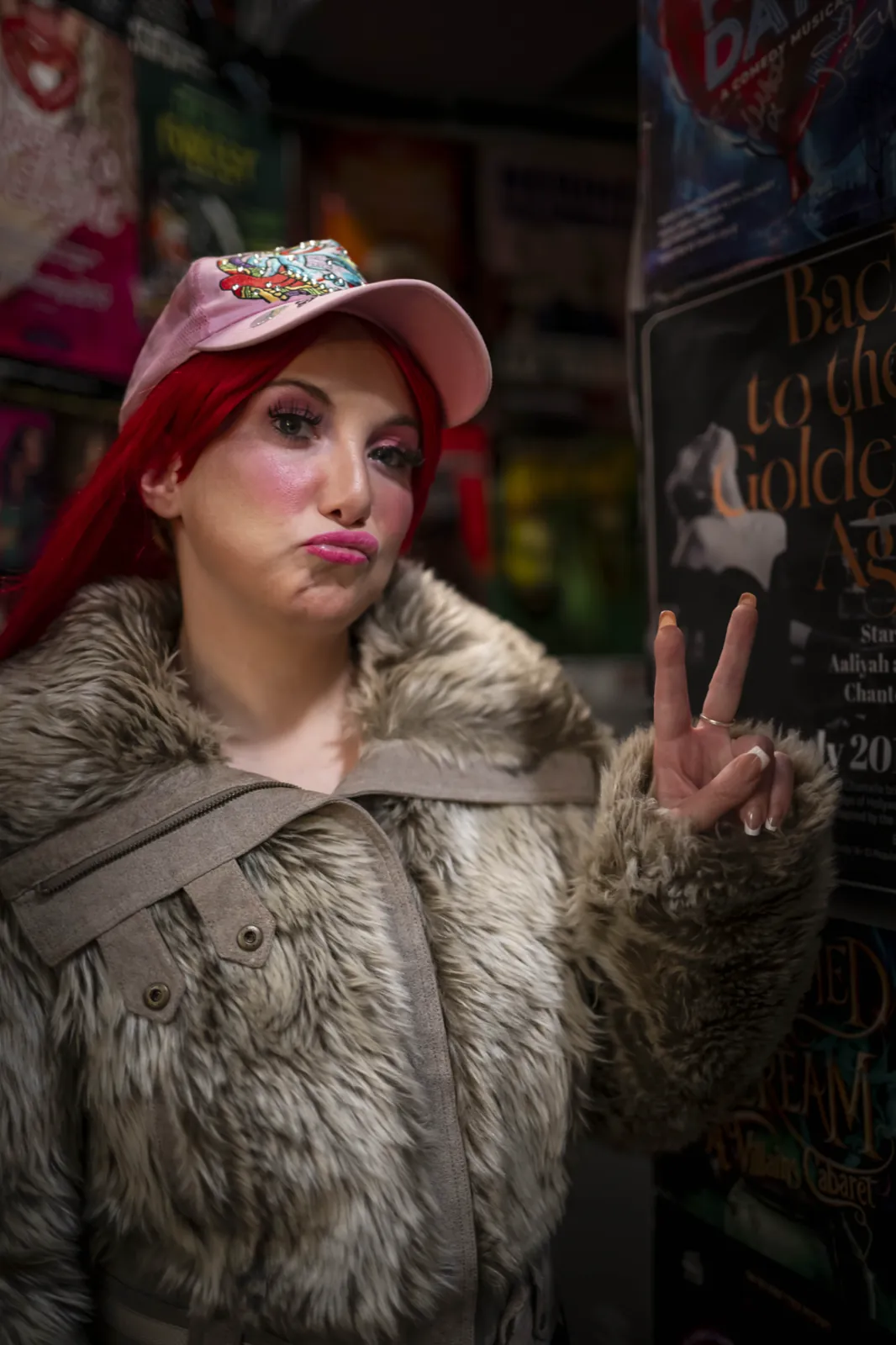 A dancer in a pink cap and fur coat in the poster-lined corridor